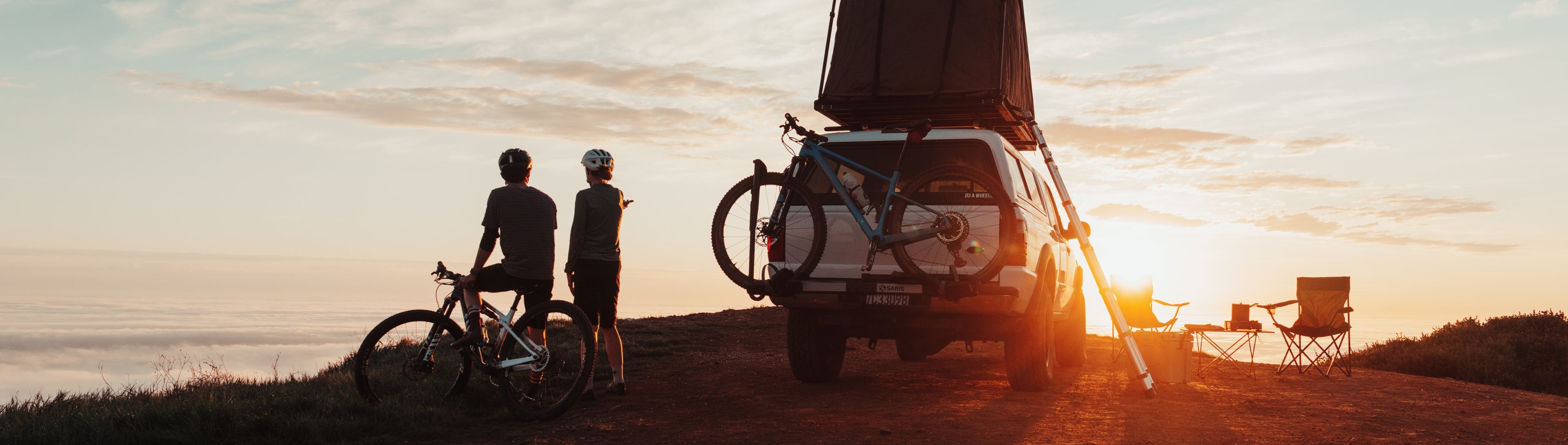 Two cyclists watching sunset from a hilltop beside a 4x4 with rooftop tent and Saris hitch rack loaded with a mountain bike