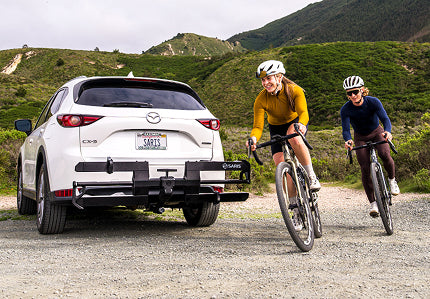 Two cyclists riding gravel bikes past a white Mazda CX-5 with a Saris hitch rack on a mountain road
