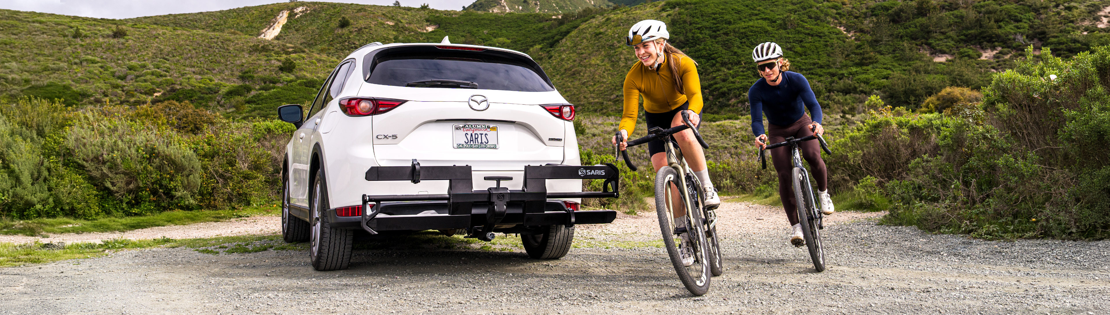 Two cyclists on gravel bikes riding past a white Mazda CX-5 with an empty Saris hitch rack on a mountain gravel road